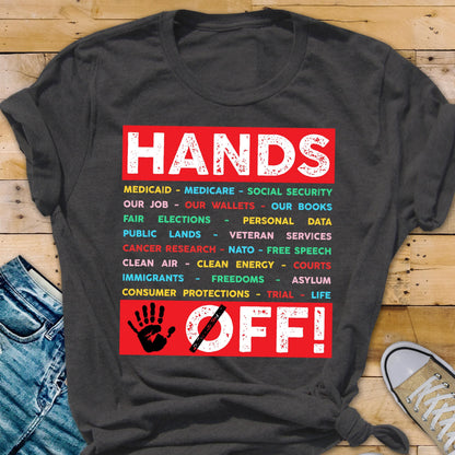 May include: A dark gray t-shirt with a red rectangle that says "HANDS OFF!" in white text. The rectangle has a red handprint with a diagonal red line through it. The rectangle also has a list of items in colorful text, including "Medicaid", "Medicare", "Social Security", "Our Job", "Our Wallets", "Our Books", "Fair Elections", "Personal Data", "Public Lands", "Veteran Services", "Cancer Research", "NATO", "Free Speech", "Clean Air", "Clean Energy", "Courts", "Immigrants", "Freedoms", "Asylum", "Consumer Pr