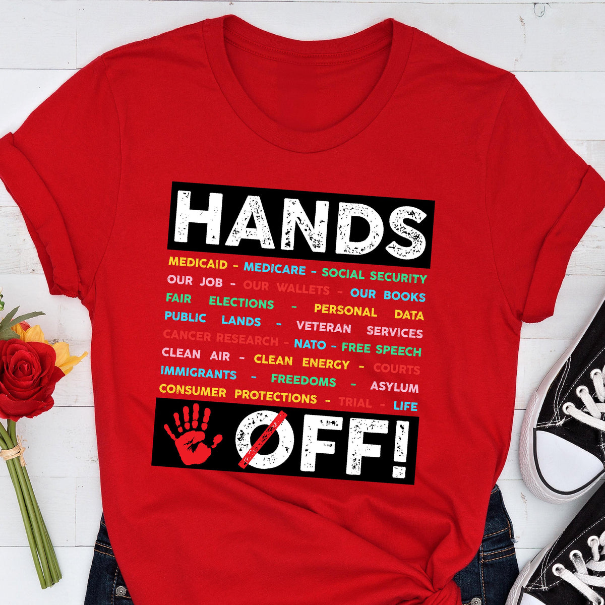May include: Red t-shirt with the text "HANDS OFF!" in black and white. The text is surrounded by a list of items in different colors, including "Medicaid", "Medicare", "Social Security", "Our Job", "Our Wallets", "Our Books", "Fair Elections", "Personal Data", "Public Lands", "Veteran Services", "Cancer Research", "NATO", "Free Speech", "Clean Air", "Clean Energy", "Courts", "Immigrants", "Freedoms", "Asylum", "Trial", "Life", and "Consumer Protections". A red hand with a diagonal line through it is below 
