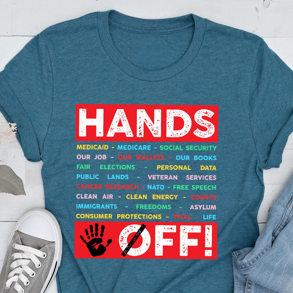 May include: A teal blue t-shirt with the text "HANDS OFF!" in red and white. The text is surrounded by a list of items in various colors, including "Medicaid", "Medicare", "Social Security", "Our Job", "Our Wallets", "Our Books", "Fair Elections", "Personal Data", "Public Lands", "Veteran Services", "Cancer Research", "NATO - Free Speech", "Clean Air", "Clean Energy", "Courts", "Immigrants", "Freedoms", "Asylum", "Life", and "Consumer Protections - Trial".