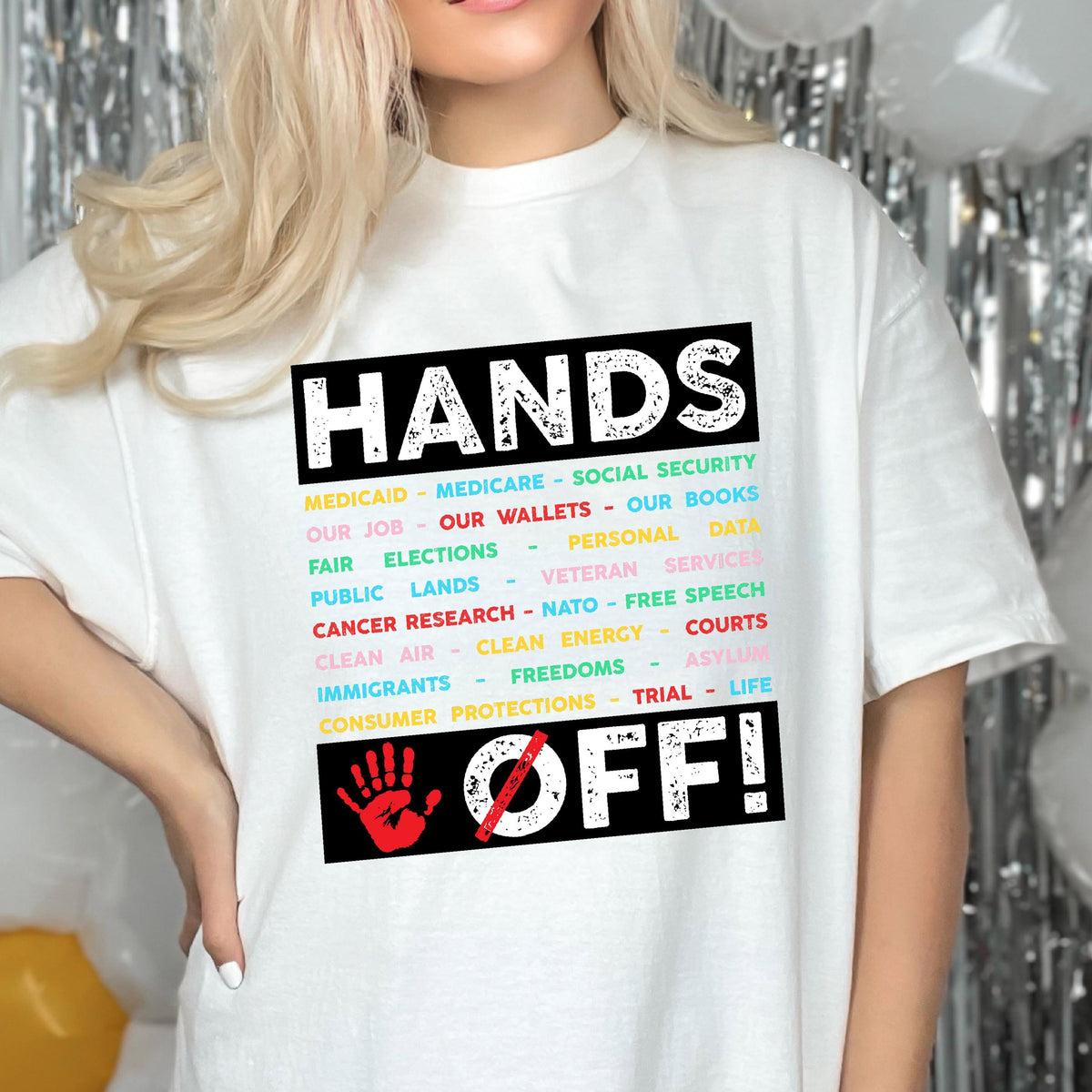 May include: White t-shirt with a black graphic that says "Hands Off!" with a list of items including Medicaid, Medicare, Social Security, our job, our wallets, our books, fair elections, personal data, public lands, veteran services, cancer research, NATO, free speech, clean air, clean energy, courts, immigrants, freedoms, asylum, consumer protections, trial, and life.
