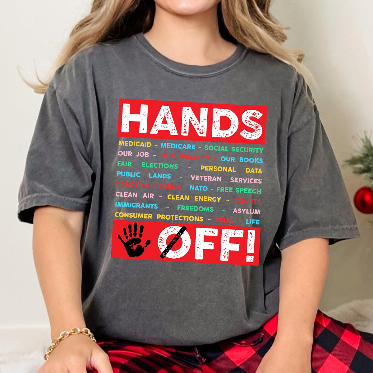 May include: A gray t-shirt with a red graphic that says "HANDS OFF!" with a list of items including "Medicaid", "Medicare", "Social Security", "Our Job", "Fair Elections", "Our Books", "Personal Data", "Public Lands", "Veteran Services", "NATO", "Free Speech", "Clean Air", "Clean Energy", "Immigrants", "Freedoms", "Asylum", "Consumer Protections", and "Life".