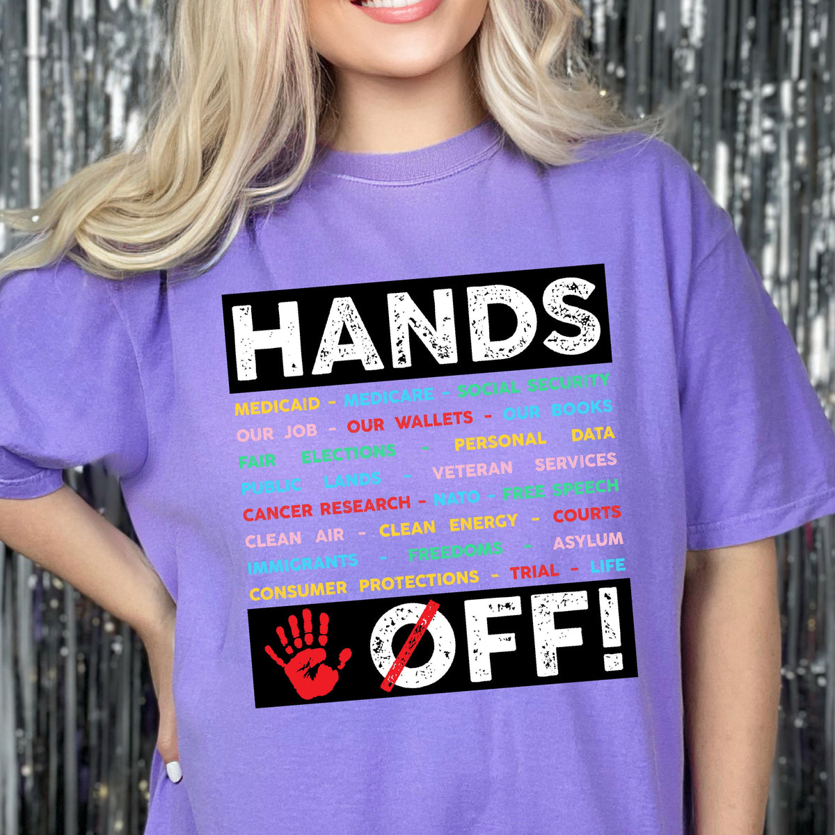 May include: Purple t-shirt with a black box that says "HANDS OFF!" in white text. The box also includes a list of items with the text "MEDICAID - MEDICINE - OUR SAFETY - OUR BOOKS - OUR JOB - OUR WALLETS - PERSONAL DATA - FAIR ELECTIONS - VETERAN SERVICES - CANCER RESEARCH - CLEAN ENERGY - COURTS - IMMIGRANTS - ASYLUM - CONSUMER PROTECTIONS - TRIAL - LIFE". A red hand with a diagonal red line through it is below the text.
