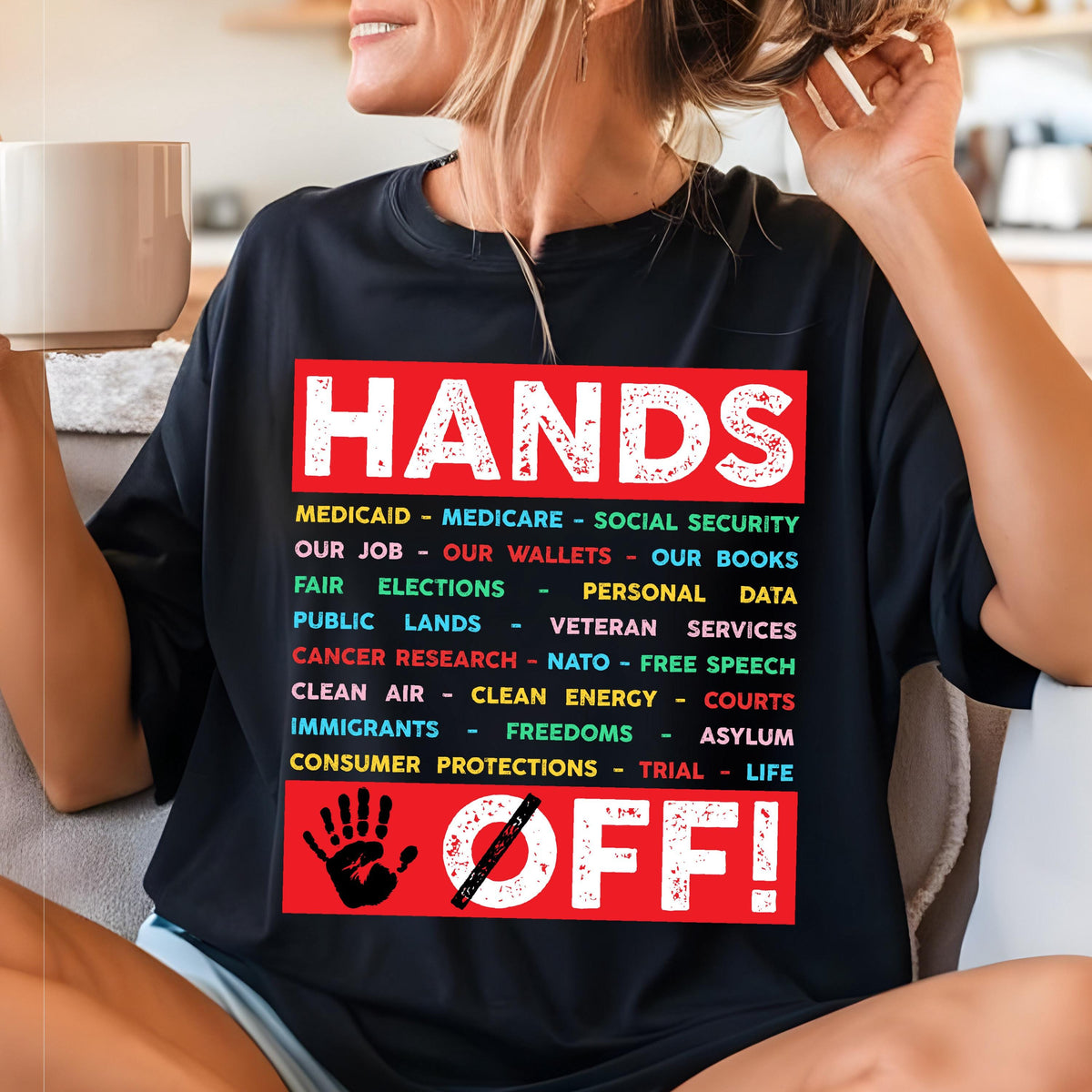 May include: Black t-shirt with a red graphic that says "Hands Off!" and lists various social programs and rights, including Medicaid, Medicare, Social Security, our jobs, our wallets, our books, fair elections, personal data, public lands, veteran services, cancer research, NATO, free speech, clean air, clean energy, courts, immigrants, freedoms, asylum, consumer protections, trial, and life.