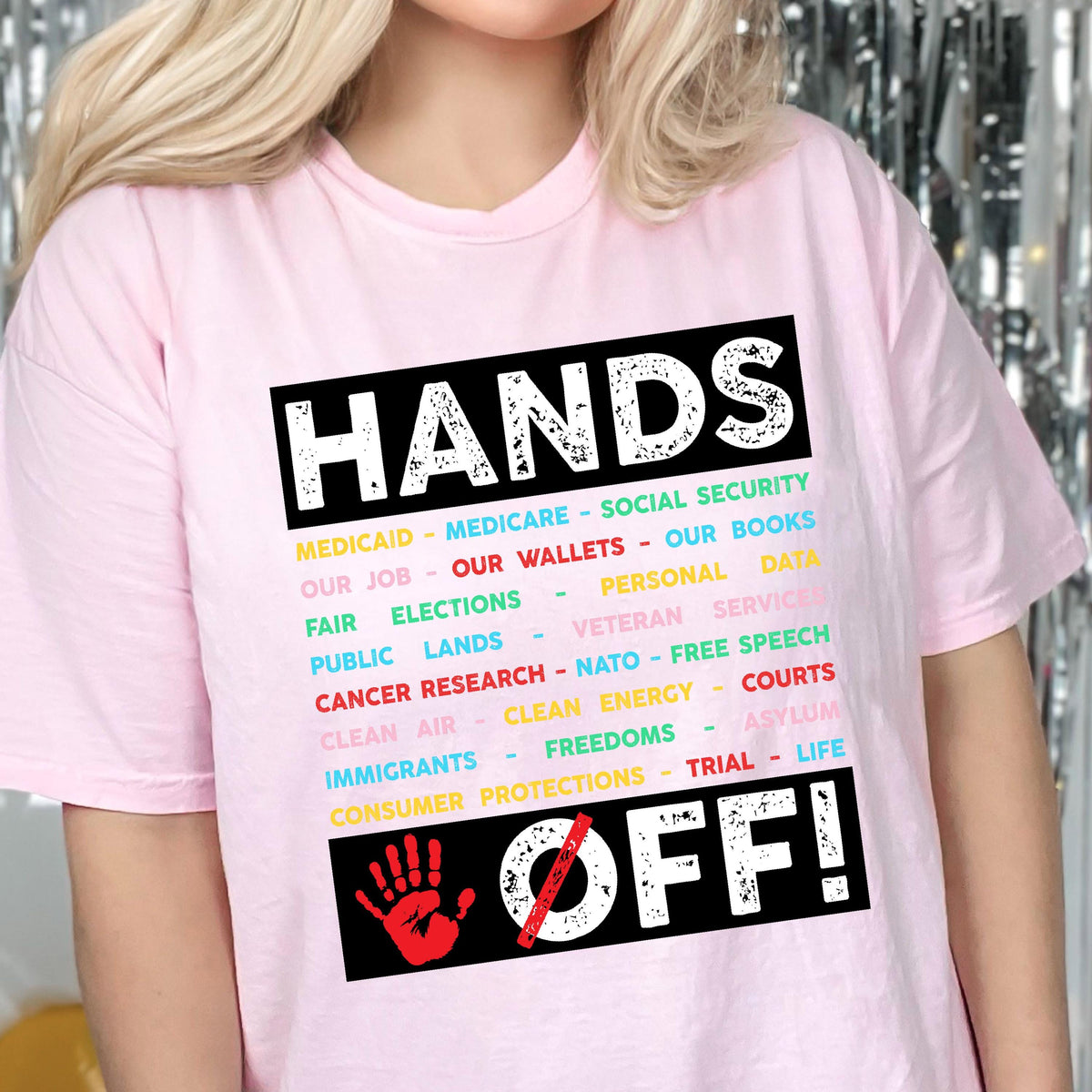 May include: Pink t-shirt with the text "HANDS OFF!" in black with a red handprint and a diagonal line through it. The text is surrounded by a list of items in various colors, including "Medicaid", "Medicare", "Social Security", "Our Job", "Our Wallets", "Our Books", "Fair Elections", "Personal Data", "Public Lands", "Veteran Service", "Cancer Research", "NATO", "Free Speech", "Clean Air", "Clean Energy", "Courts", "Immigrants", "Freedoms", "Asylum", "Consumer Protections", "Trial", and "Life".