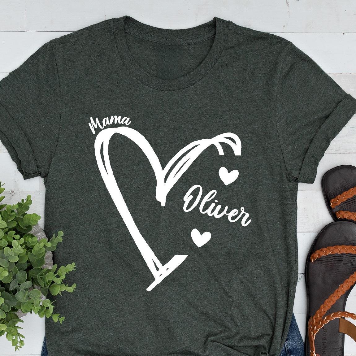 May include: A dark green t-shirt with a white heart design. The heart is divided into two sections, one labeled "Mama" and the other labeled "Oliver". There are three small white hearts inside the heart design.