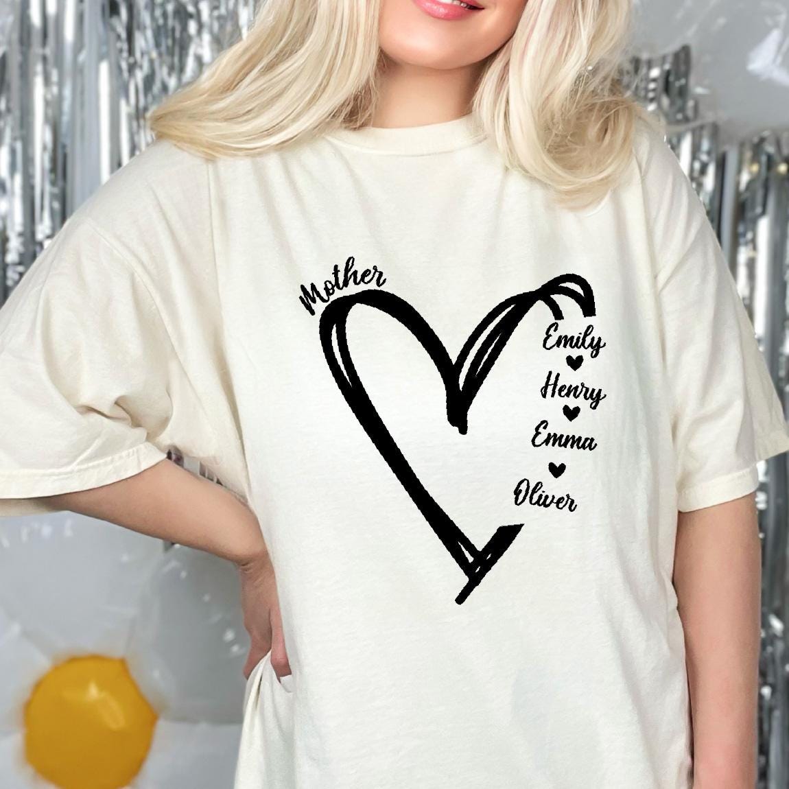 May include: A cream-colored short-sleeve t-shirt featuring a black hand-drawn heart graphic. The word Mother is printed above the heart, and the names Emily, Henry, Emma, and Oliver are listed vertically inside the heart, separated by small hearts.