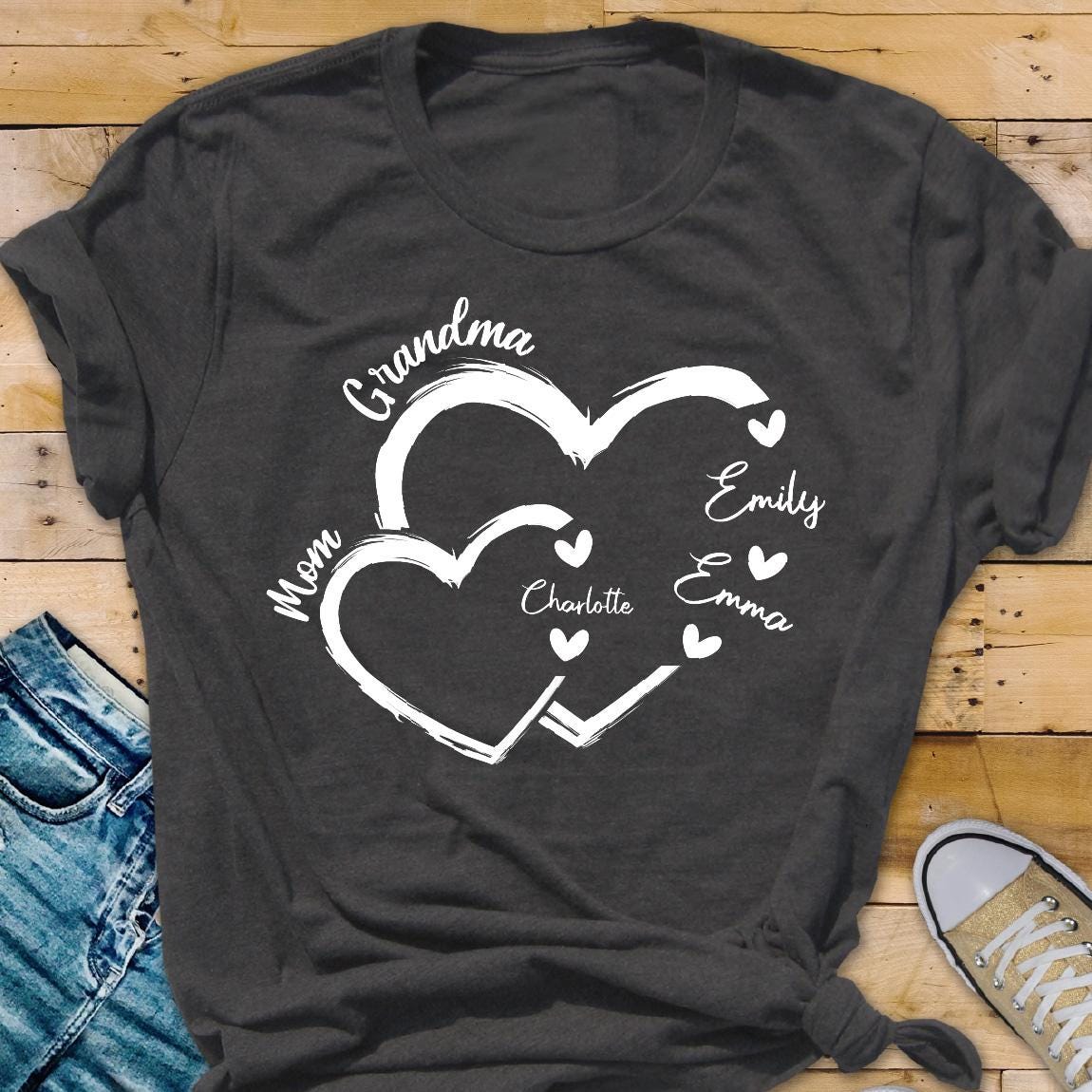 May include: Gray t-shirt with a white design of two overlapping hearts. The text "Grandma" is above the hearts, and the text "Mom" is to the left of the hearts. Inside the hearts are the names "Emily", "Emma", and "Charlotte".