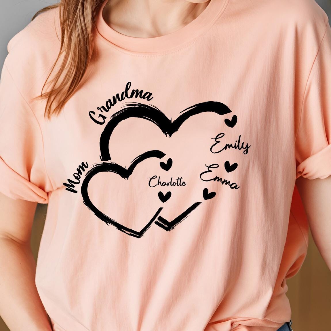 May include: A peach-colored t-shirt with a black design of two interlocking hearts. The hearts have the words "Grandma", "Mom", "Charlotte", "Emily", and "Emma" written on them.