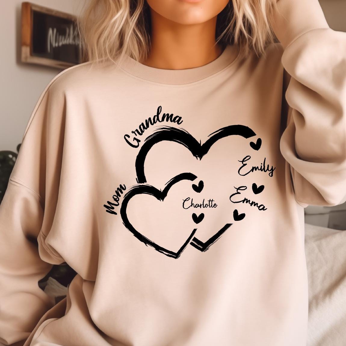 May include: A light beige sweatshirt with a black graphic design of two intertwined hearts. The text "Grandma" is above the hearts, and the text "Mom" is to the left of the hearts. The names "Charlotte", "Emily", and "Emma" are written inside the hearts.
