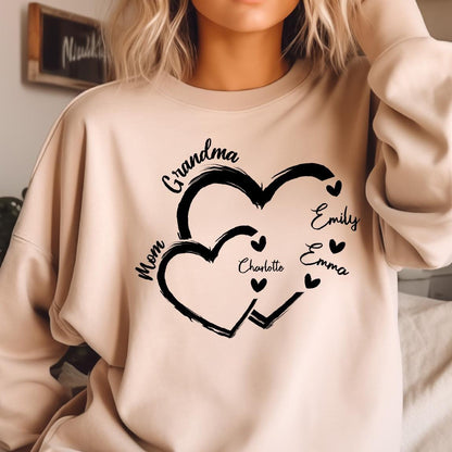 May include: A light beige sweatshirt with a black graphic design of two intertwined hearts. The text "Grandma" is above the hearts, and the text "Mom" is to the left of the hearts. The names "Charlotte", "Emily", and "Emma" are written inside the hearts.