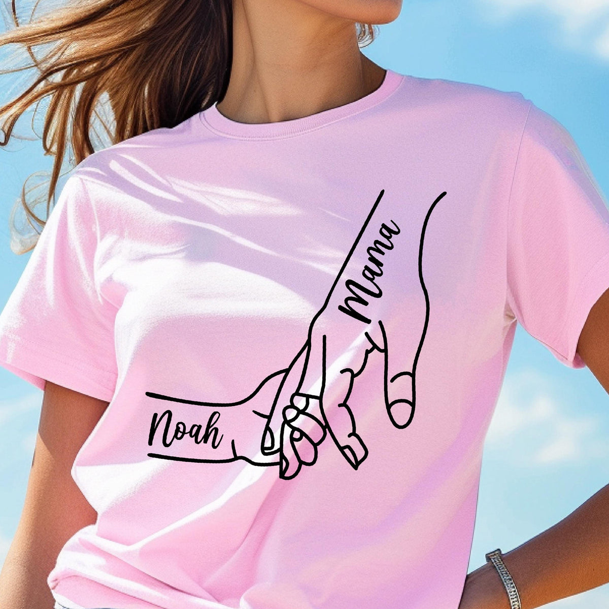 May include: A light pink t-shirt featuring a black line drawing of two hands holding each other. The word "Mama" is written vertically on one hand, and the name "Noah" is written on the other hand.