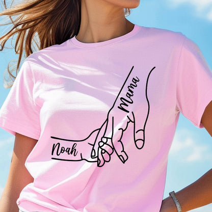 May include: A light pink t-shirt featuring a black line drawing of two hands holding each other. The word "Mama" is written vertically on one hand, and the name "Noah" is written on the other hand.