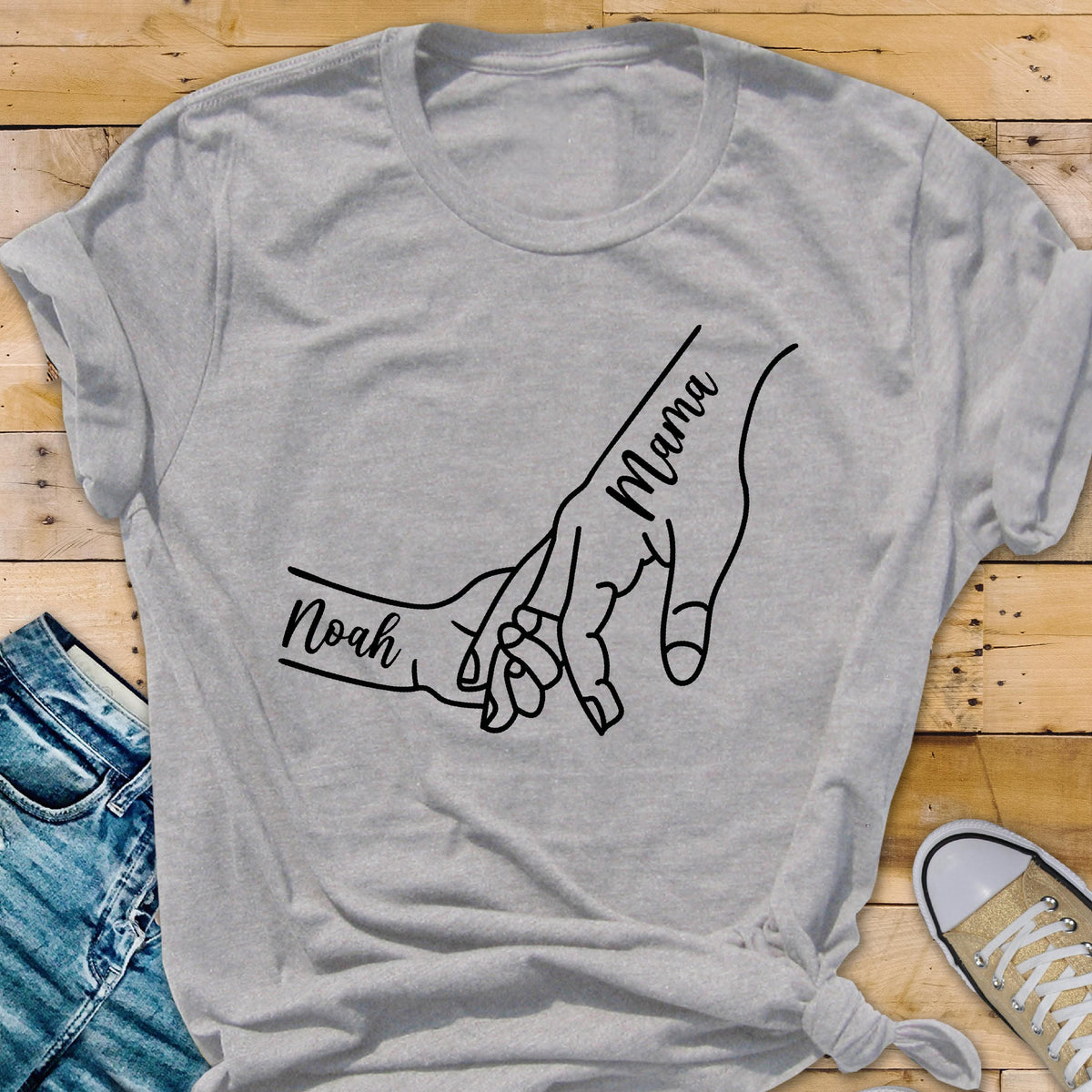 May include: A gray t-shirt featuring a black line drawing of two hands holding each other. The word "Mama" is written on one hand, and the name "Noah" is written on the other. The shirt is made of a soft material.