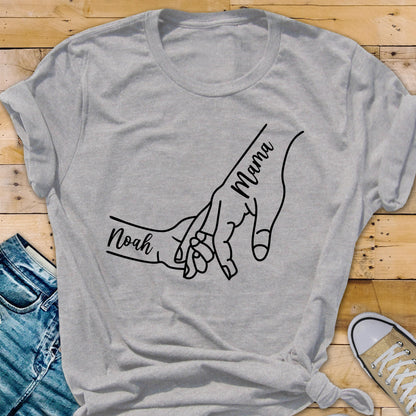 May include: A gray t-shirt featuring a black line drawing of two hands holding each other. The word "Mama" is written on one hand, and the name "Noah" is written on the other. The shirt is made of a soft material.