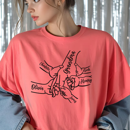 May include: A coral-colored t-shirt featuring a black line-art graphic of multiple hands holding onto one central hand. The central hand is labeled Grandma, while the surrounding hands are labeled with the names Charlotte, Noah, Henry, and James.