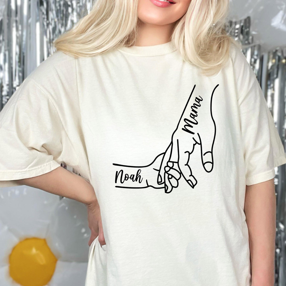 May include: An off-white short-sleeve t-shirt featuring a black line art graphic of two hands holding each other. The larger hand is labeled with the word Mama, and the smaller hand is labeled with the name Noah in a cursive script font.