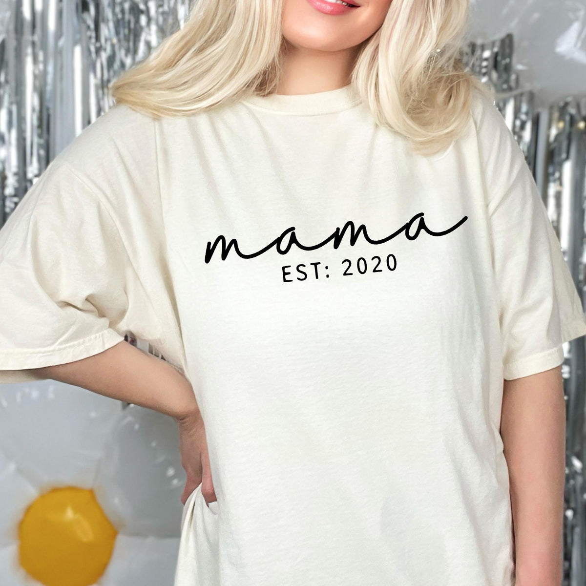 May include: An off-white short-sleeve t-shirt featuring the black text "mama" in a cursive script font, with the text "EST: 2020" printed in a simple sans-serif font directly underneath. The shirt has a crew neckline and a relaxed fit.