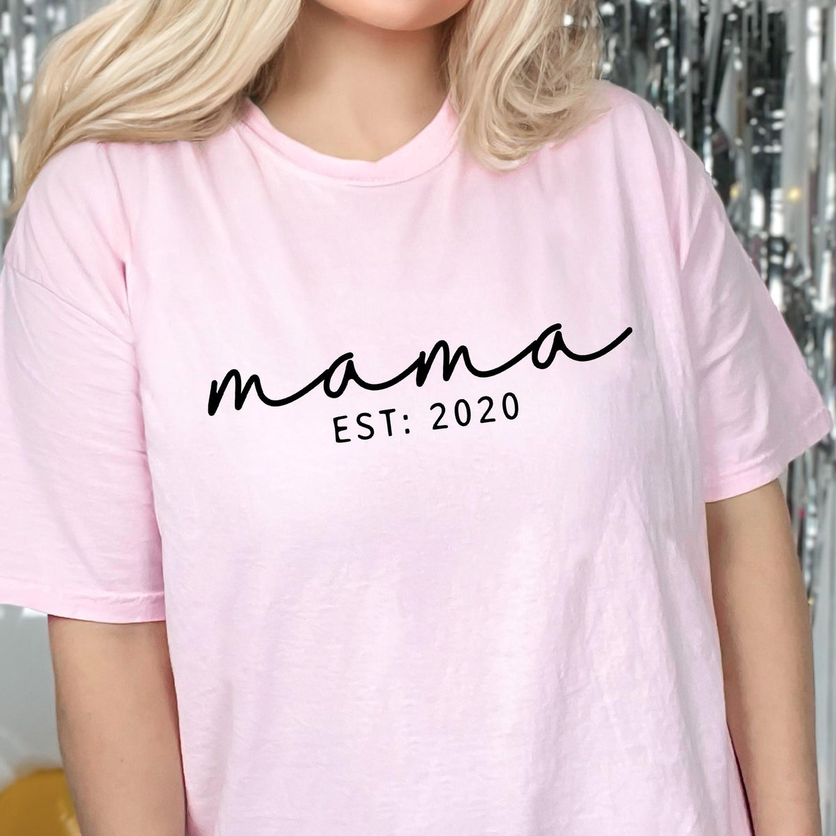 May include: A light pink t-shirt with the word "mama" in a black cursive font. Below the word "mama" is the text "EST: 2020" in a smaller, black, sans-serif font. The shirt has short sleeves.