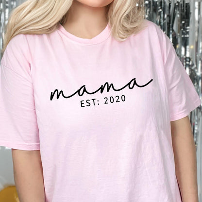 May include: A light pink t-shirt with the word "mama" in a black cursive font. Below the word "mama" is the text "EST: 2020" in a smaller, black, sans-serif font. The shirt has short sleeves.