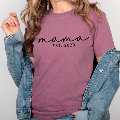 May include: A mauve short-sleeved t-shirt featuring the black text "mama" in a cursive script font, with the text "EST: 2020" printed in a simple sans-serif font directly underneath. The shirt is worn under a blue denim jacket.