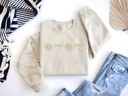 May include: A beige sweatshirt with a simple design of five daisies with bows on a light beige background.
