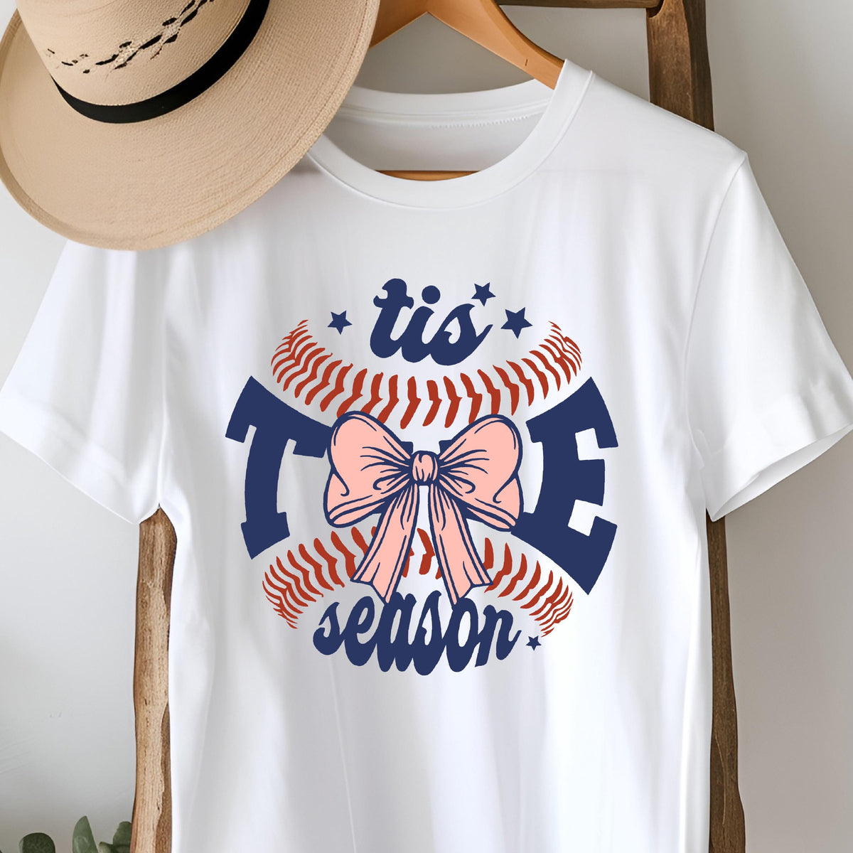 May include: White t-shirt with a blue and red baseball graphic and a pink bow. The graphic says "tis the season".