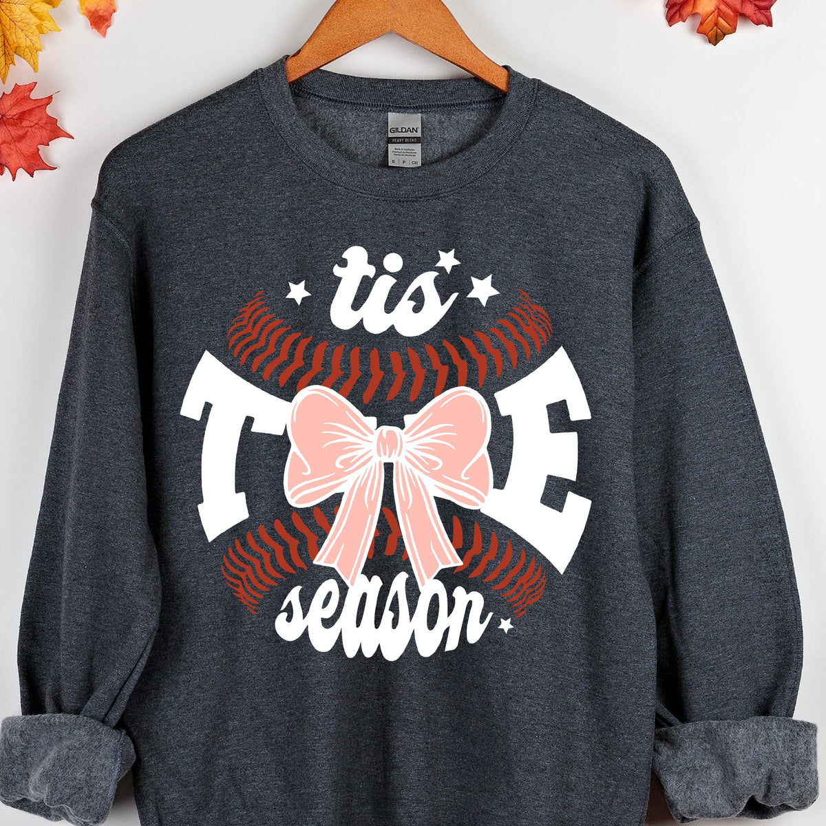 May include: A dark gray sweatshirt with a white baseball graphic and the text "'tis the season" in white. A pink bow is in the center of the baseball graphic.