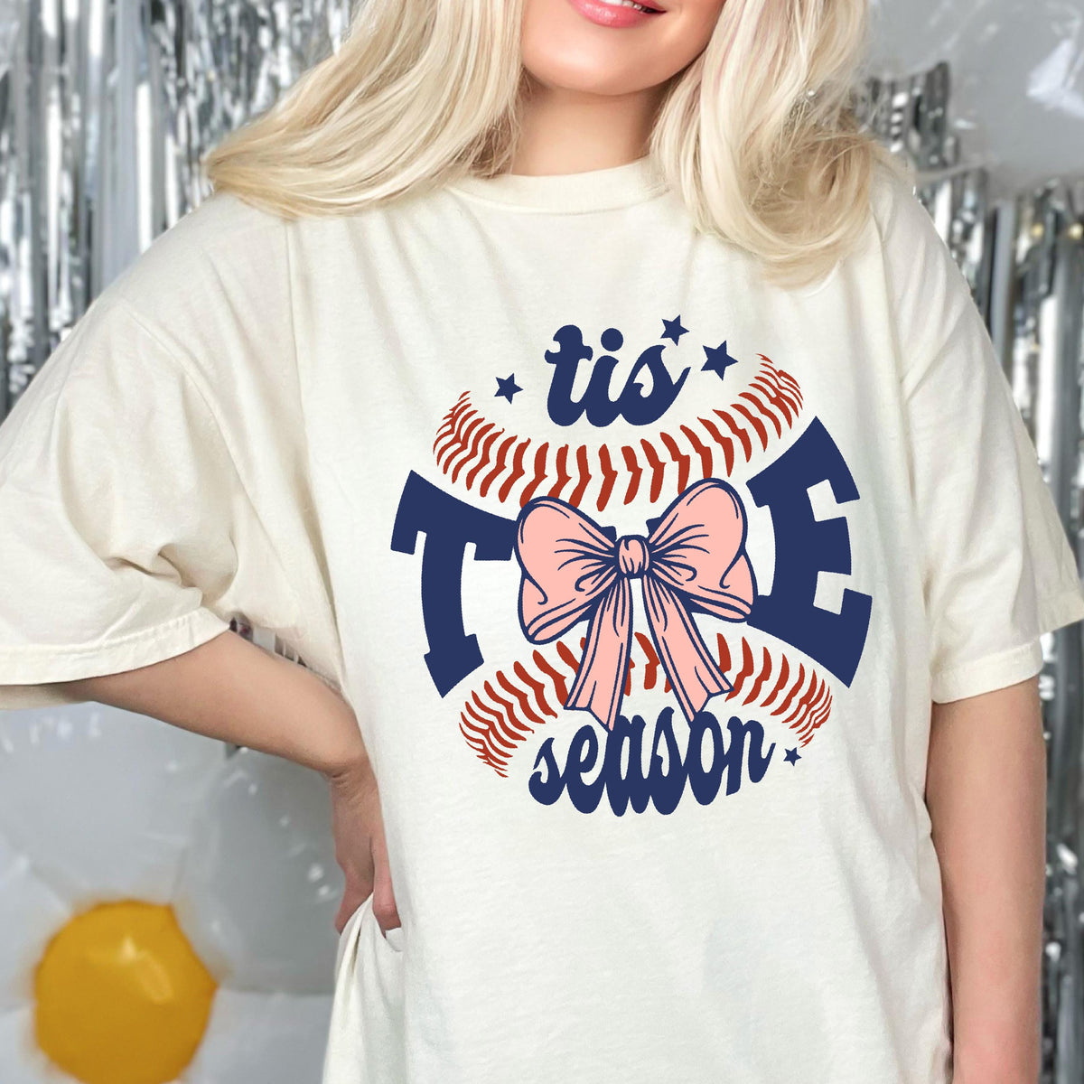 May include: A white t-shirt with a baseball graphic and the text "tis the season" in navy blue. A pink bow is in the center of the baseball graphic.