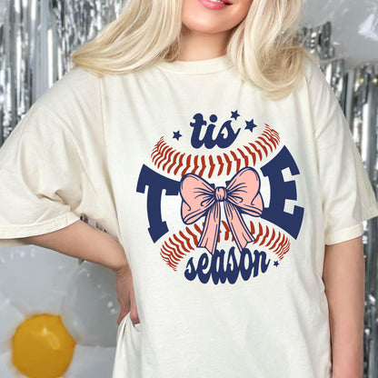 May include: A white t-shirt with a baseball graphic and the text "tis the season" in navy blue. A pink bow is in the center of the baseball graphic.