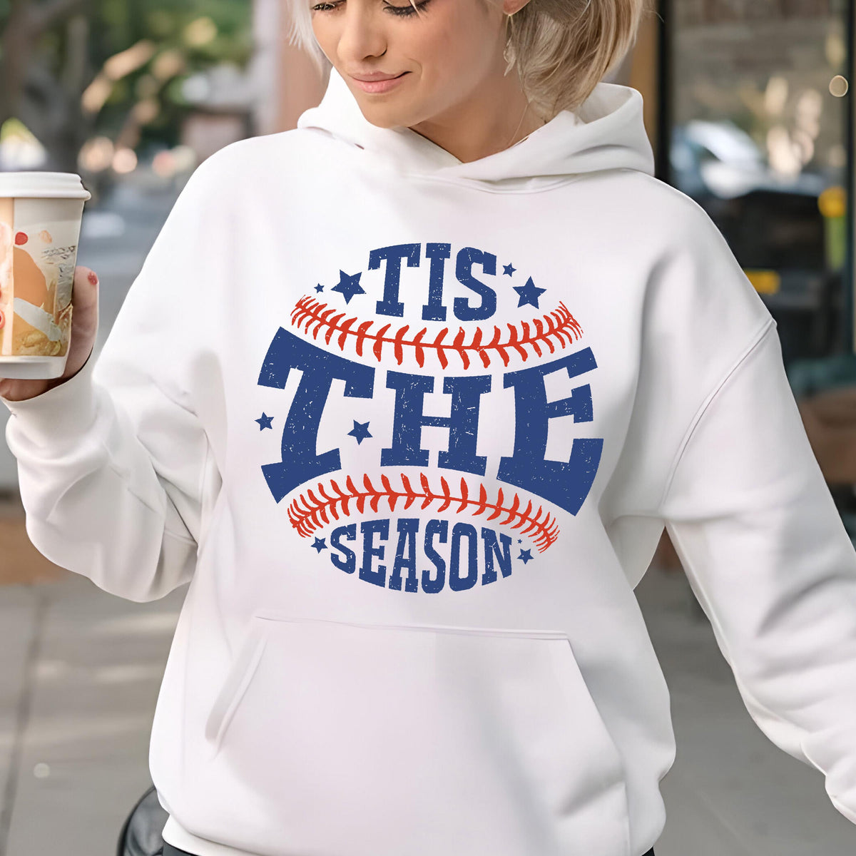 May include: A white hooded sweatshirt featuring a graphic design of a baseball with red stitching and blue text that reads TIS THE SEASON. The design includes small blue stars and has a distressed texture. The hoodie has long sleeves and a front pocket.