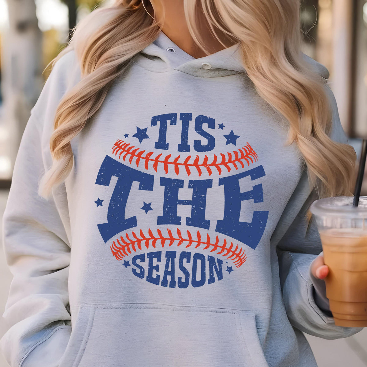 May include: A light blue hooded sweatshirt featuring a graphic design of a baseball with red stitching. The text 'TIS THE SEASON' is printed in blue block letters across the center, accented by small blue stars surrounding the baseball graphic.