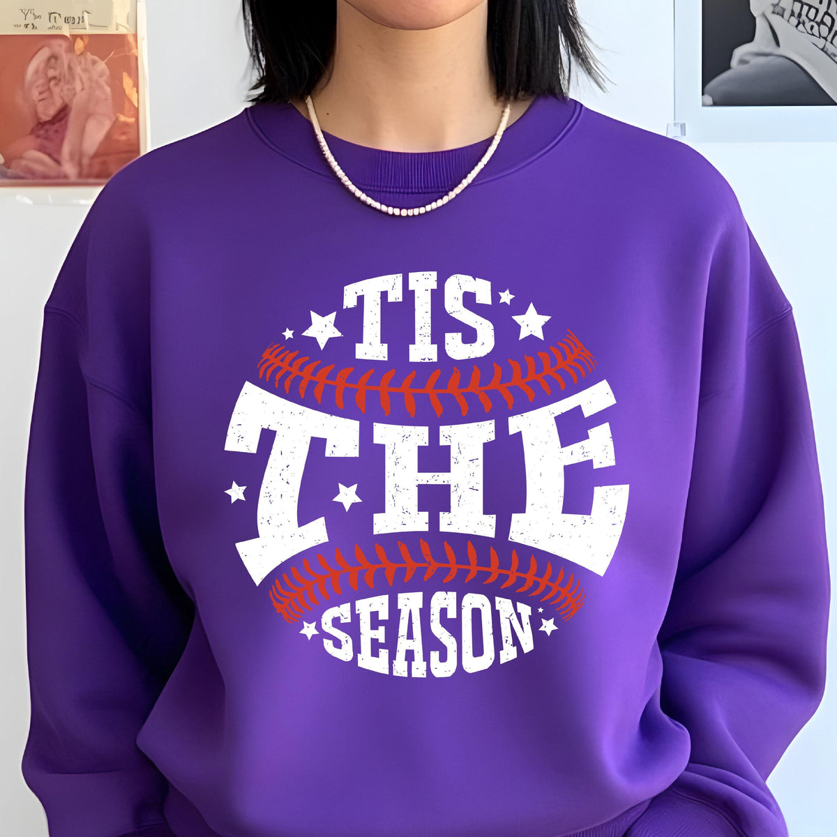 May include: A purple crewneck sweatshirt featuring a graphic design of a baseball with red stitching. The white distressed text reads 'TIS THE SEASON' across the center, accented by small white stars positioned around the baseball graphic.