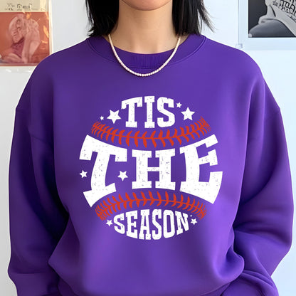 May include: A purple crewneck sweatshirt featuring a graphic design of a baseball with red stitching. The white distressed text reads 'TIS THE SEASON' across the center, accented by small white stars positioned around the baseball graphic.
