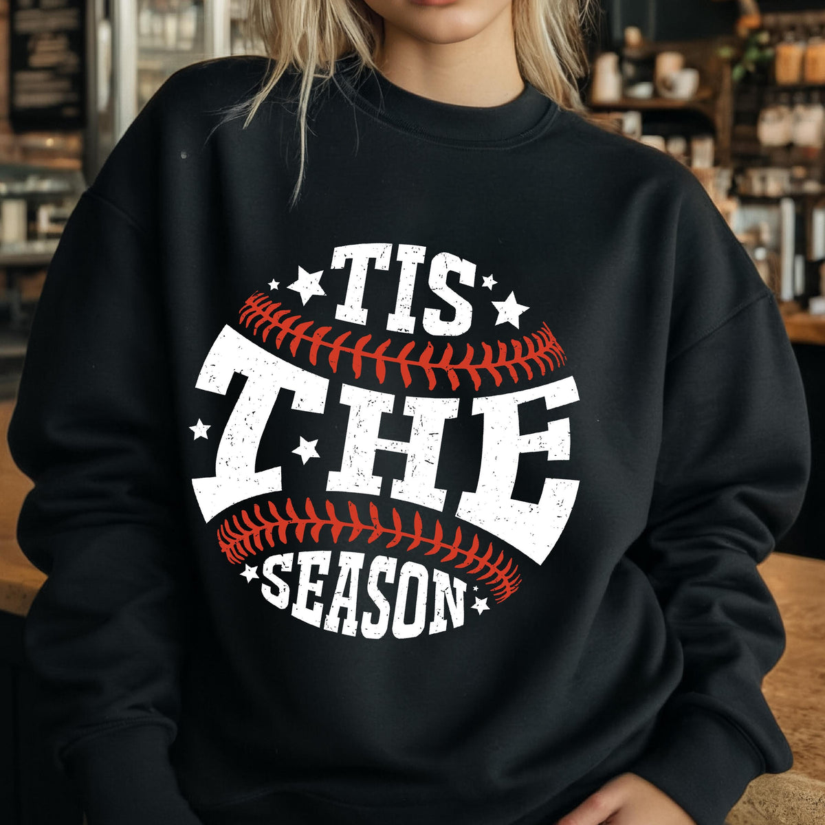 May include: A black crewneck sweatshirt featuring a graphic design of a baseball with red stitching. The white distressed text reads TIS THE SEASON, arranged in a circular pattern around the baseball, accented with small white stars.