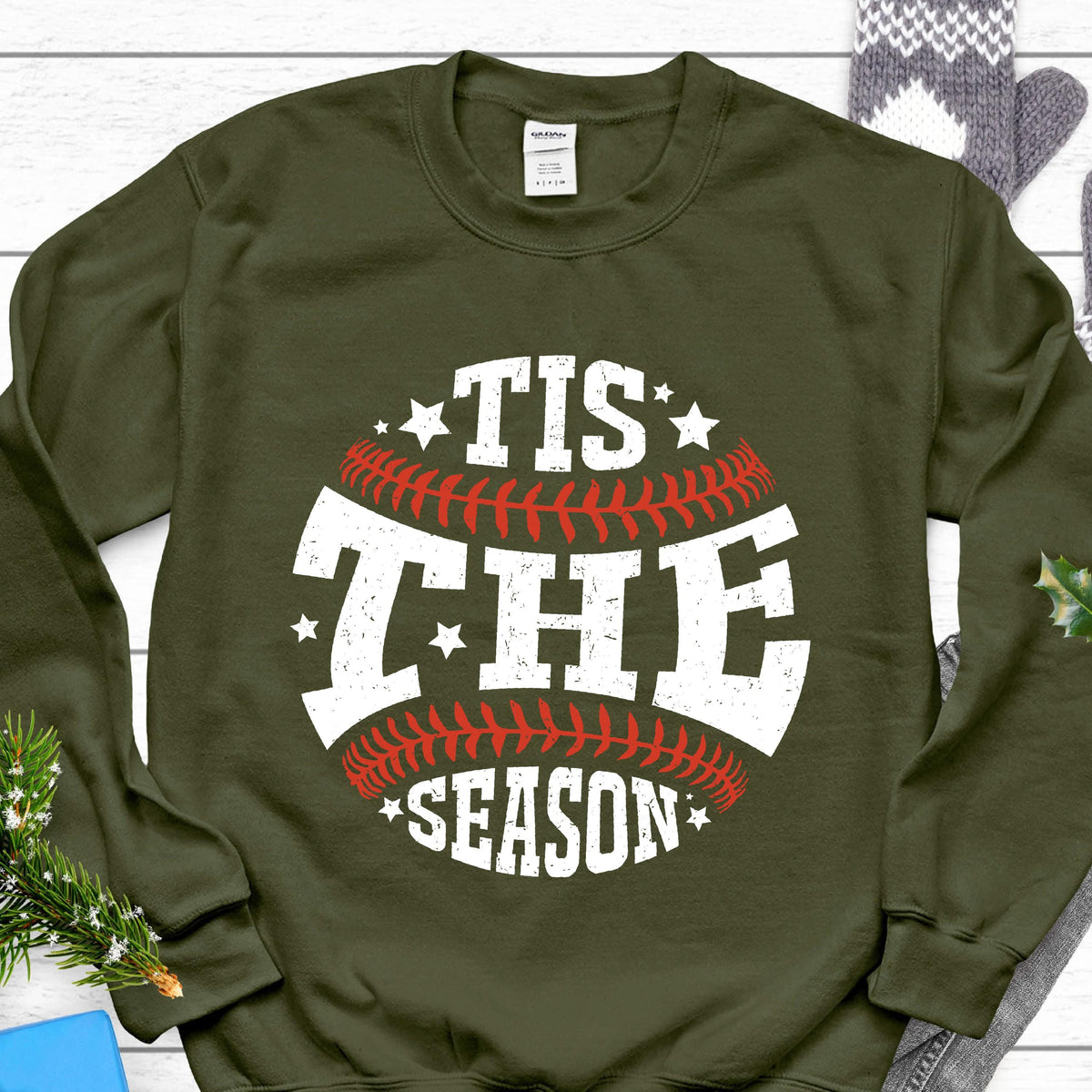 May include: An olive green crewneck sweatshirt featuring a white graphic design of a baseball with red stitching. The text "TIS THE SEASON" is printed in a distressed white font across the center, accented by small white stars around the design.