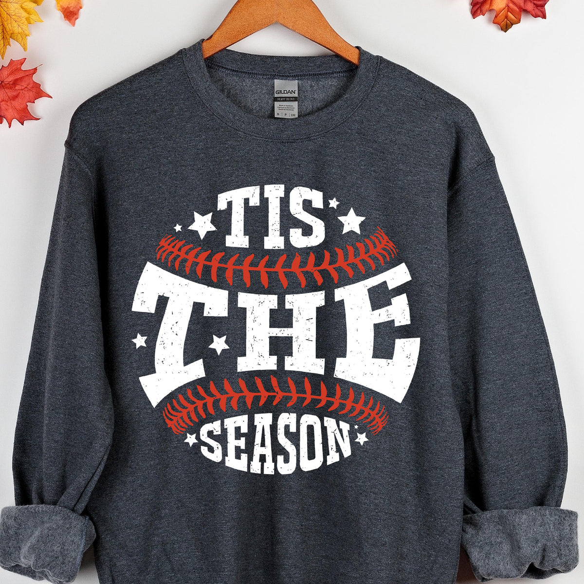 May include: A dark heather gray crewneck sweatshirt featuring a graphic of a baseball with red stitching. The white distressed text reads 'TIS THE SEASON' arranged in a circular pattern around the baseball, accented by small white stars.
