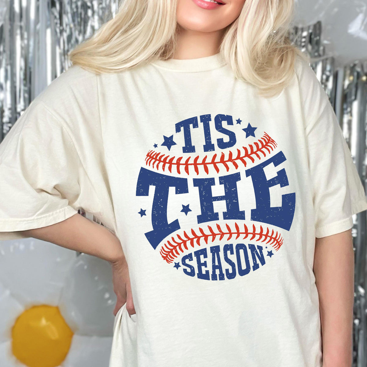 May include: A white t-shirt with a blue and red baseball graphic. The graphic says "Tis the Season" with stars around the baseball.