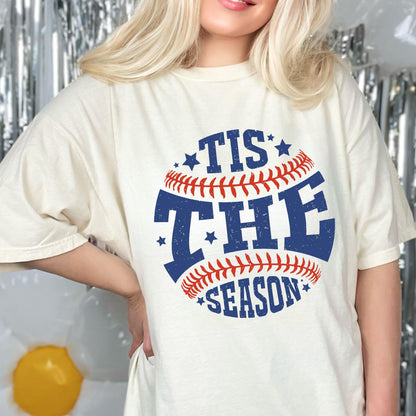 May include: A white t-shirt with a blue and red baseball graphic. The graphic says "Tis the Season" with stars around the baseball.