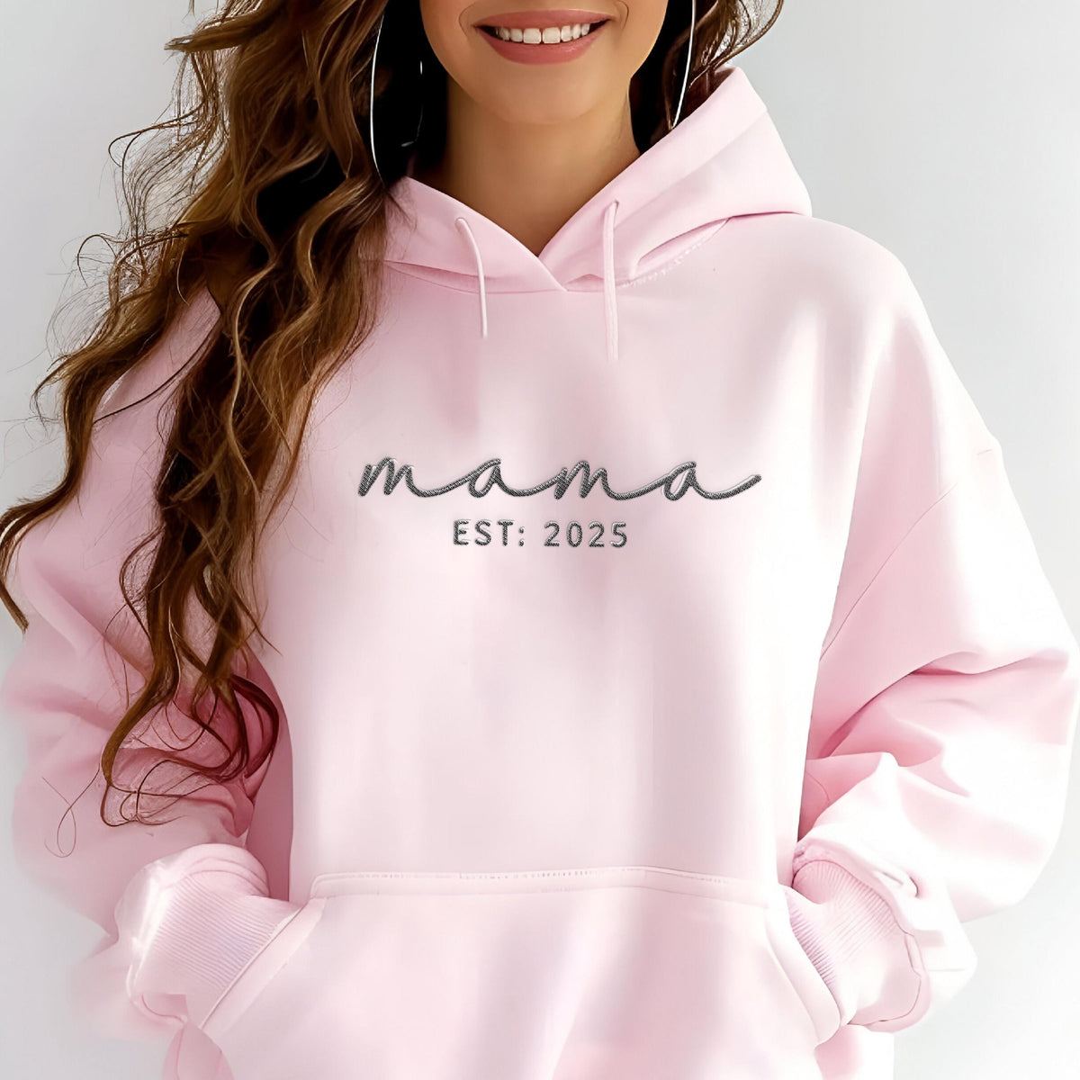 May include: A light pink hooded sweatshirt featuring the word mama in a dark gray cursive script font, with the text EST: 2025 printed in a simple sans-serif font directly underneath. The garment includes a front kangaroo pocket and drawstrings.
