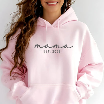 May include: A light pink hooded sweatshirt featuring the word mama in a dark gray cursive script font, with the text EST: 2025 printed in a simple sans-serif font directly underneath. The garment includes a front kangaroo pocket and drawstrings.