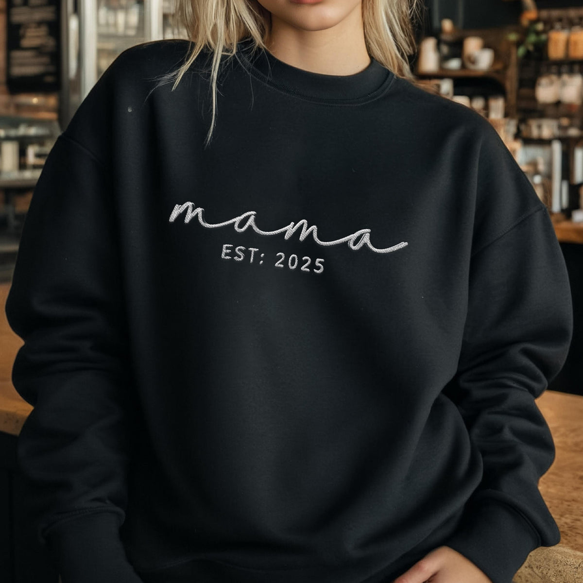 May include: A black long-sleeve crewneck sweatshirt featuring white embroidery on the chest. The text reads "mama" in a cursive script font, with the text "EST: 2025" embroidered in a clean, sans-serif font directly underneath the word mama.