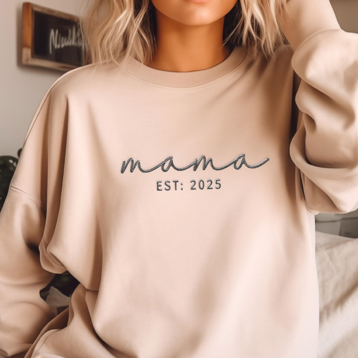 May include: A beige crewneck sweatshirt featuring the word mama in a dark gray cursive script font, with the text EST: 2025 embroidered in a simple sans-serif font directly underneath the cursive lettering on the center of the chest.