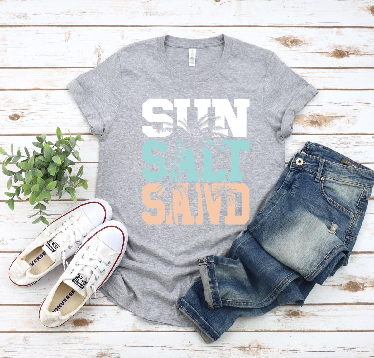 May include: A gray t-shirt with the text "SUN SALT SAND" in white, teal, and peach. The text is layered with palm tree graphics. The shirt is laid out on a white wood background with a pair of white sneakers and blue jeans.