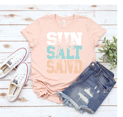 May include: A pink t-shirt with the text "SUN SALT SAND" in white, blue, and orange. The text is layered with a palm tree graphic in the middle.