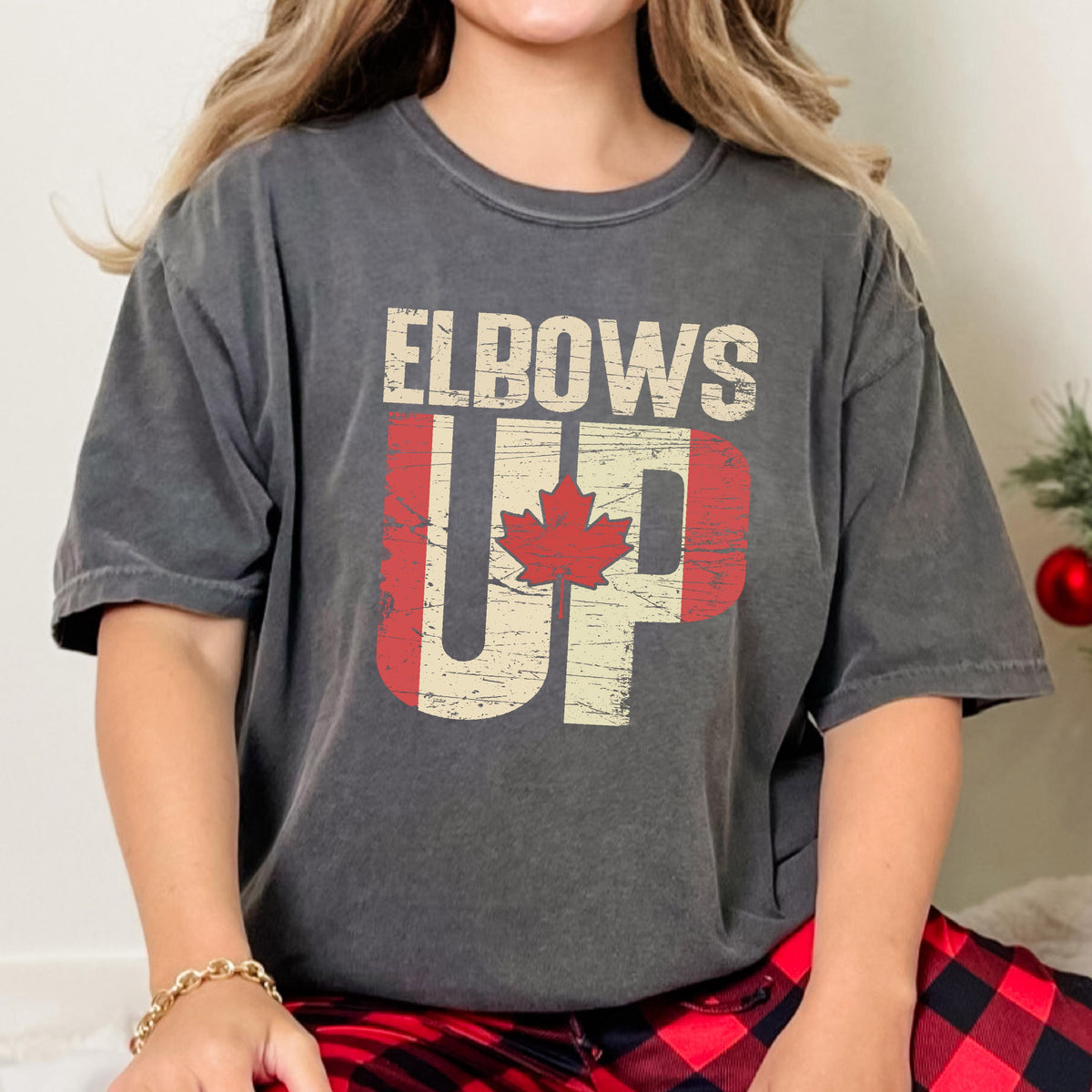 May include: A gray t-shirt with the text "Elbows Up" in white letters with a Canadian flag design behind the text. The flag is red and white with a red maple leaf.