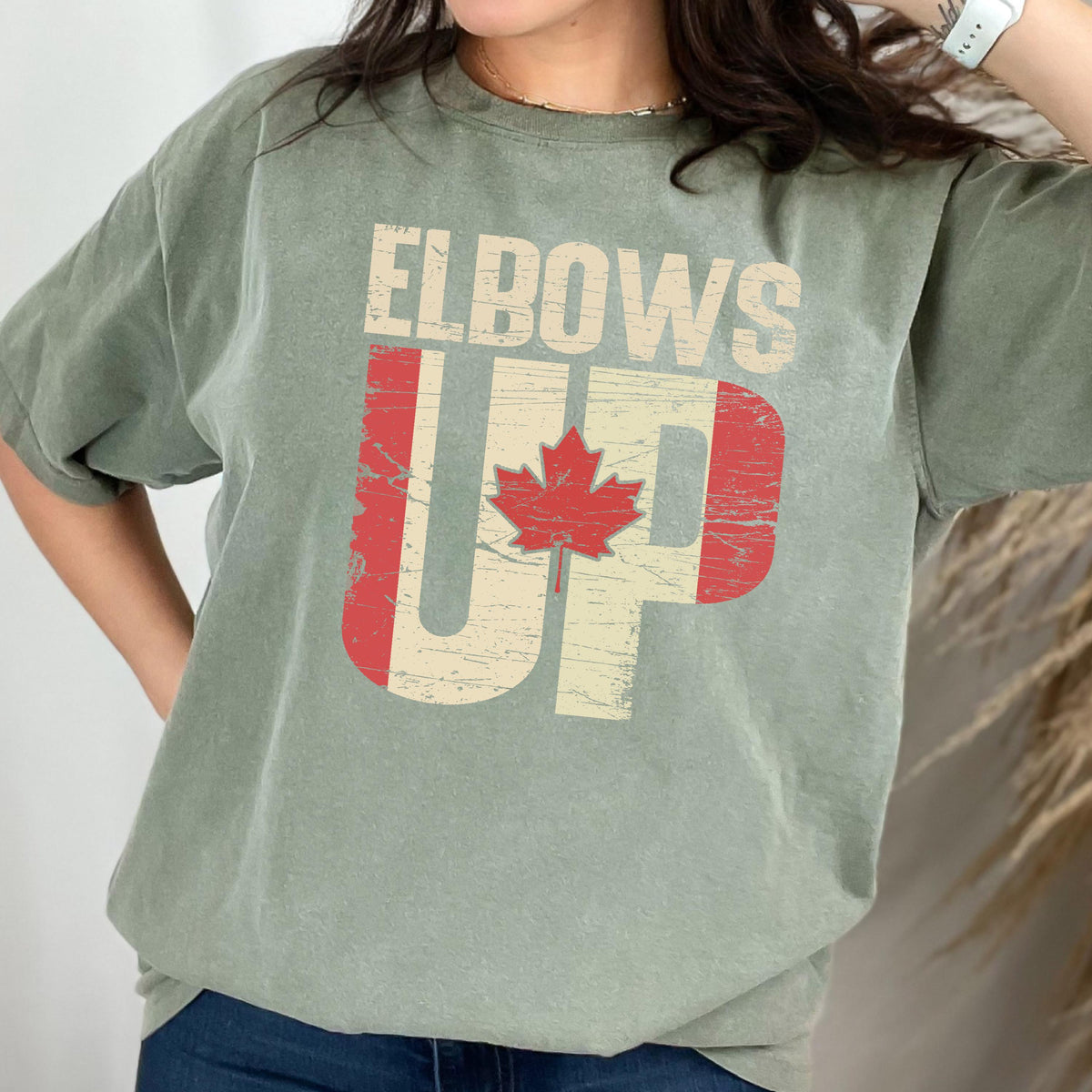 May include: A green t-shirt with the text "Elbows Up" printed on it in white. The text is overlaid with a distressed Canadian flag design.