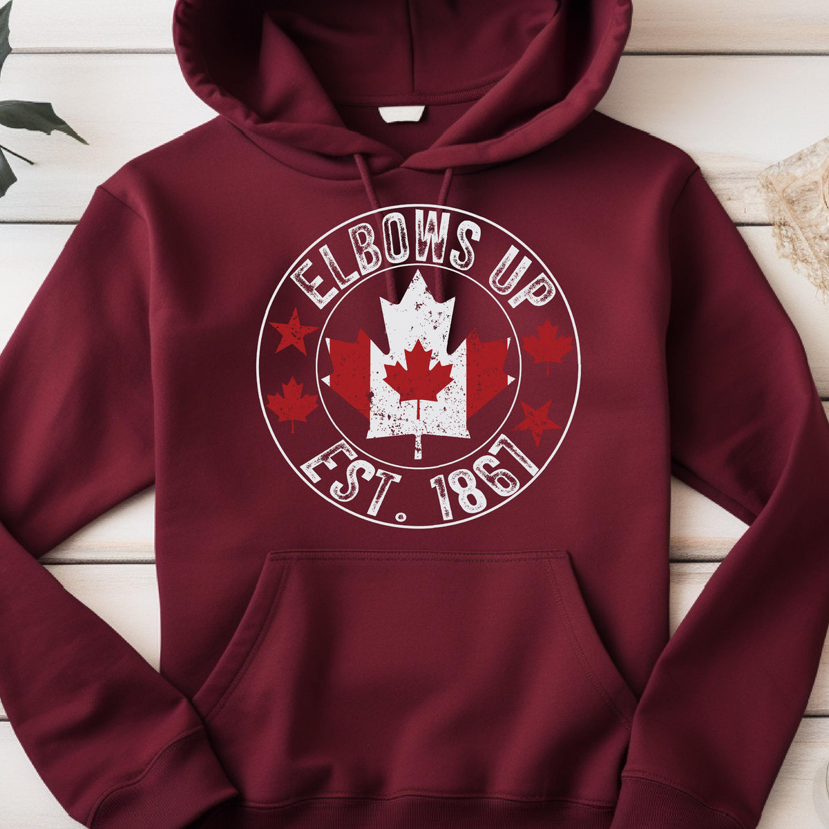 May include: A maroon hooded sweatshirt featuring a white circular graphic on the chest. The design includes the text "ELBOWS UP" and "EST. 1867" surrounding a distressed white maple leaf icon with a smaller red maple leaf centered inside.
