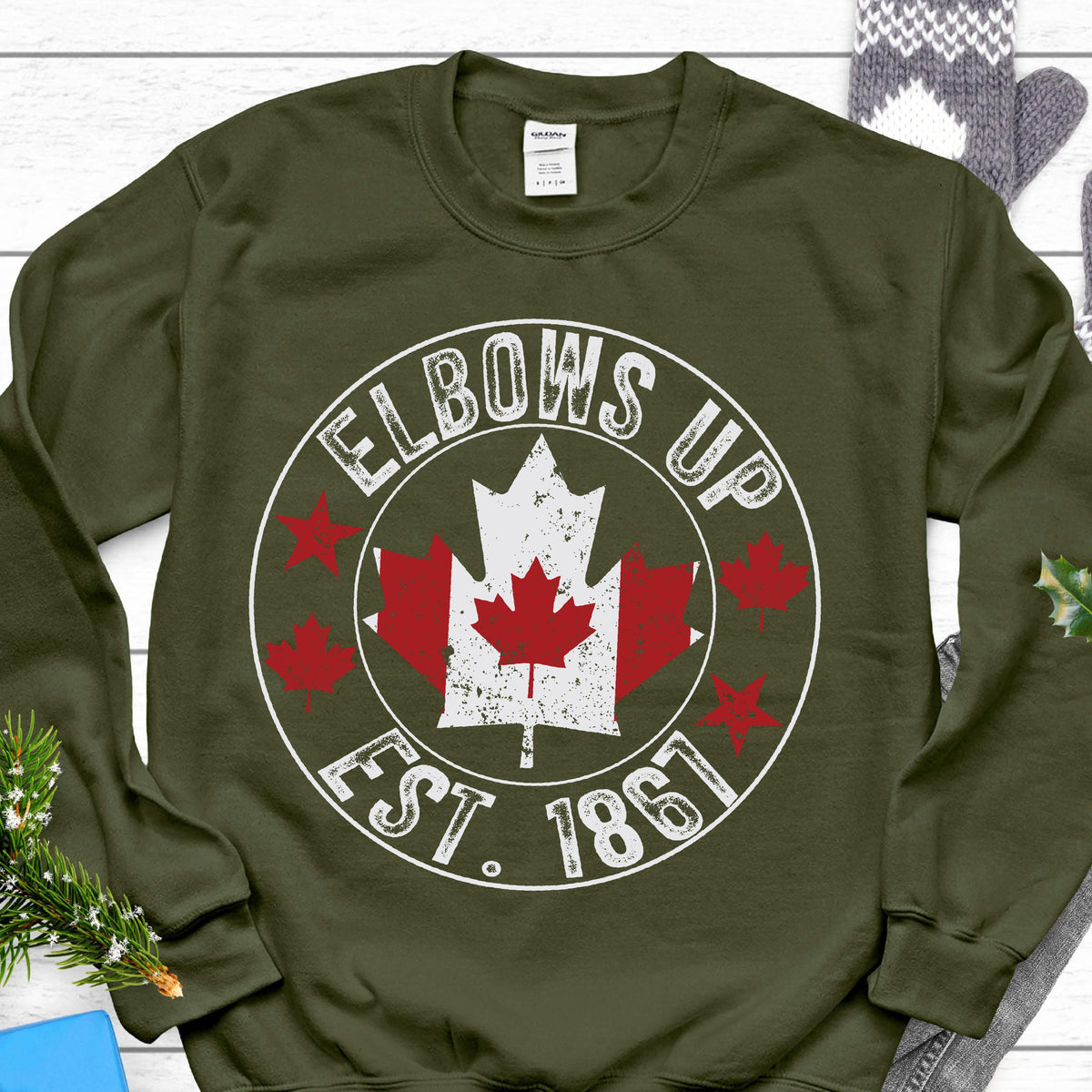 May include: An olive green crewneck sweatshirt featuring a white circular graphic with the text ELBOWS UP and EST. 1867. The design includes a central Canadian maple leaf in red and white, surrounded by four smaller red maple leaf icons.