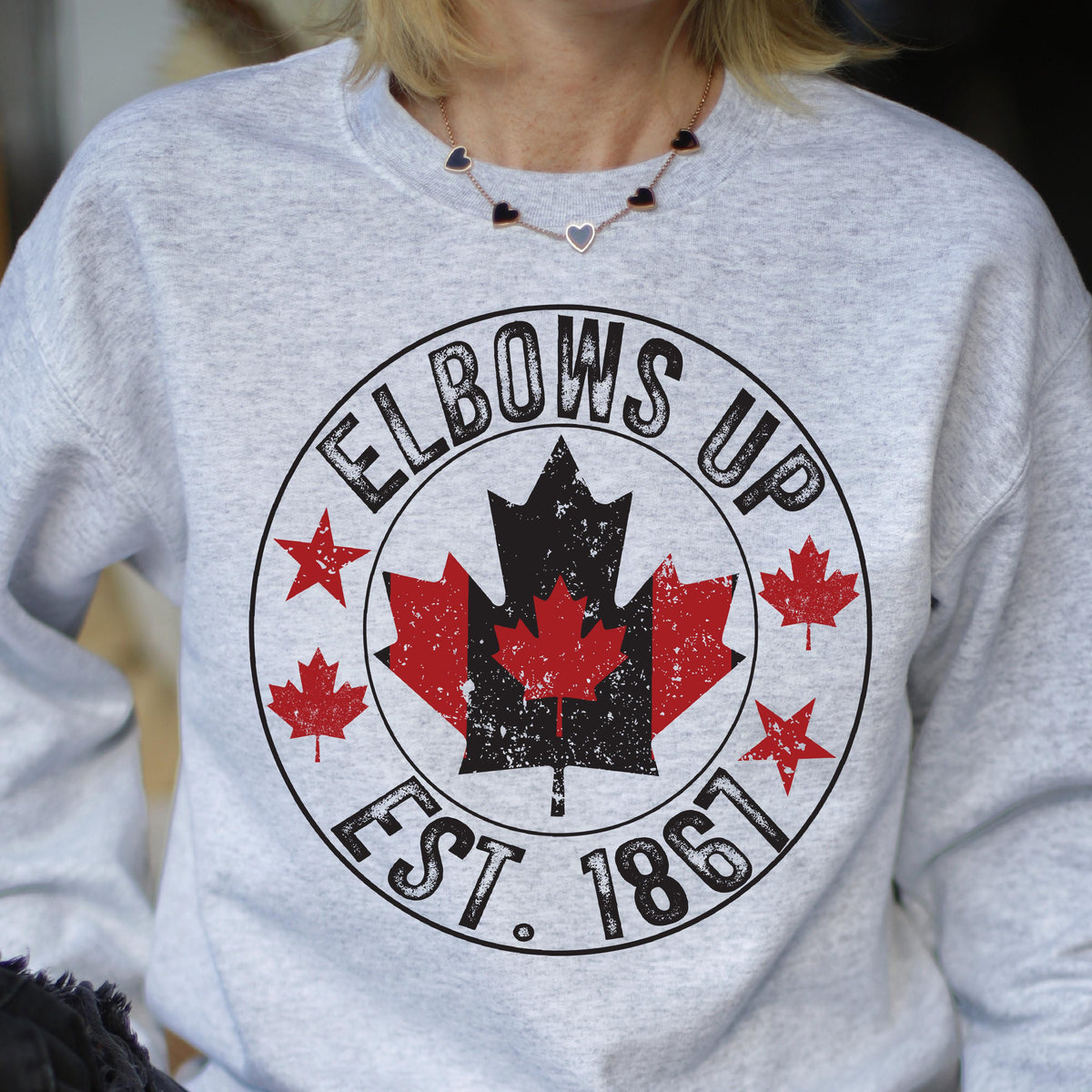May include: A light gray crewneck sweatshirt featuring a circular graphic with the text ELBOWS UP and EST. 1867. The design includes a large central maple leaf in red and black, flanked by smaller red maple leaves and stars in a distressed print style.