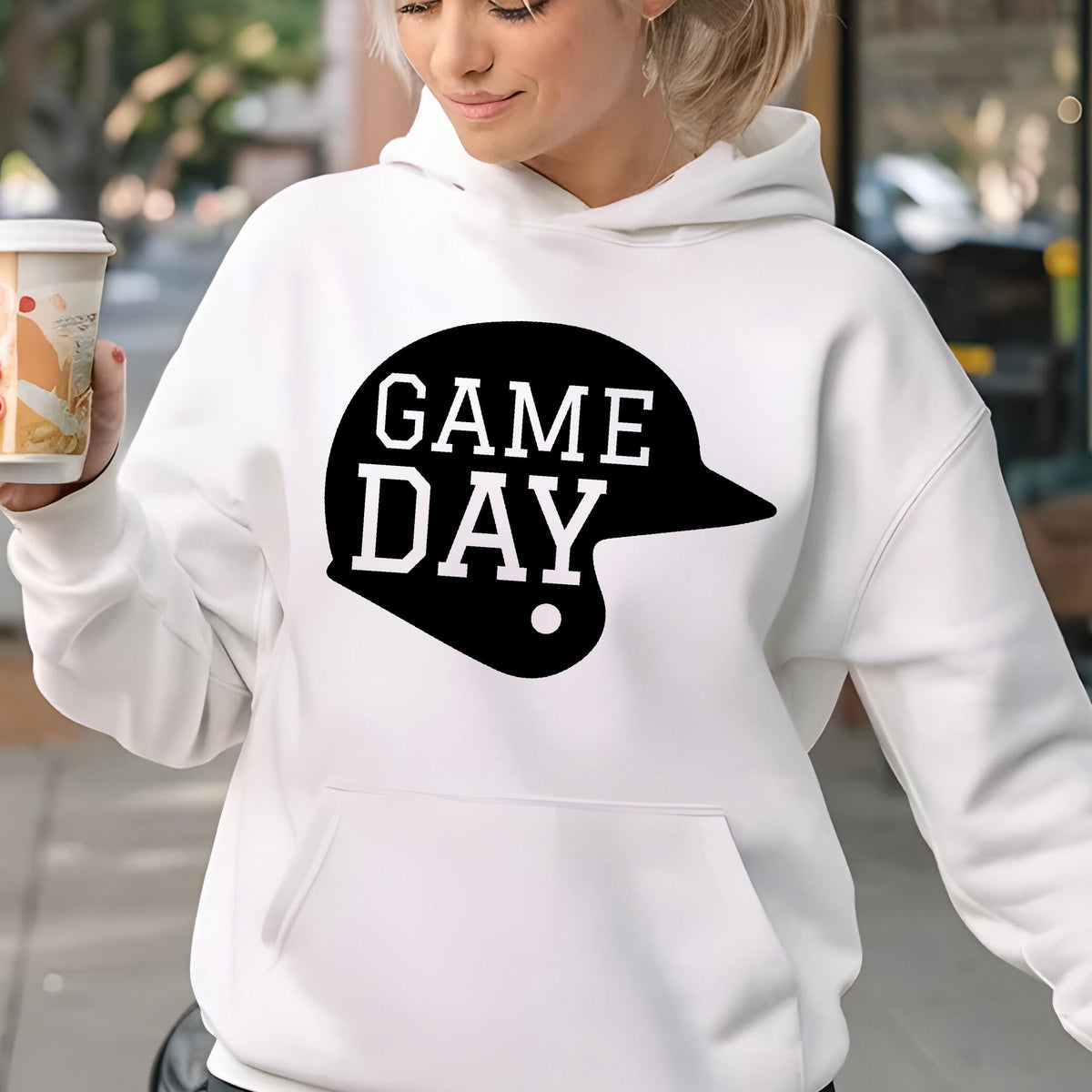 May include: A white long-sleeve hooded sweatshirt featuring a black graphic of a baseball helmet with the text GAME DAY printed in white block letters across the center of the helmet shape. The hoodie includes a front kangaroo pocket.